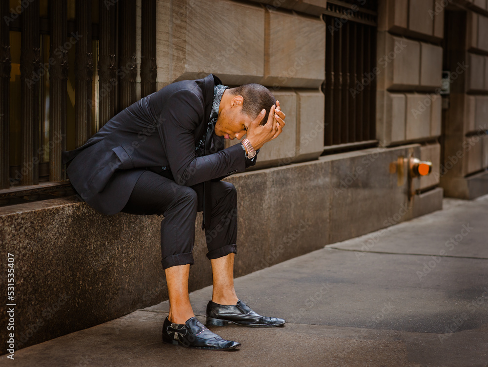 Young African American man, wearing black suit, tie, leather shoes, wristwatch, sitting on window on street in New York City, bending back, hands covering head, tired, sad, thinking..