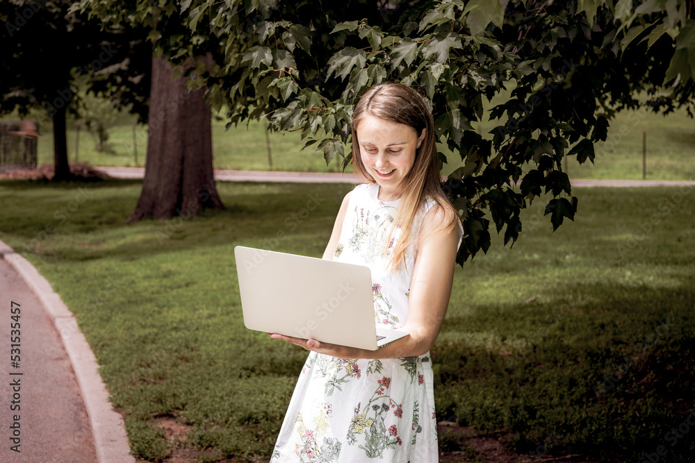 Young college female student, wearing flower patterned white dress, standing on green lawn by trees on campus in New York City, reading, working on laptop computer. .