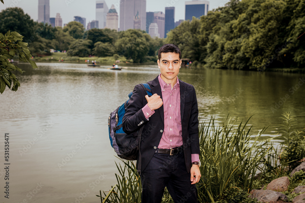 Young college male student, wearing black blazer, red, white patterned under shirt, carrying shoulder bag, standing by lake in New York City, looking forward..