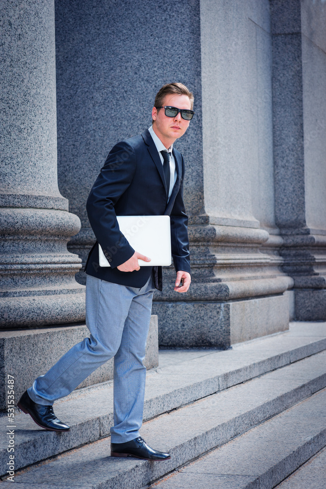 Young Man, wearing black blazer, necktie, gray pants, leather shoes, sunglasses, carrying laptop computer, walking down stairs from office building in New York City..