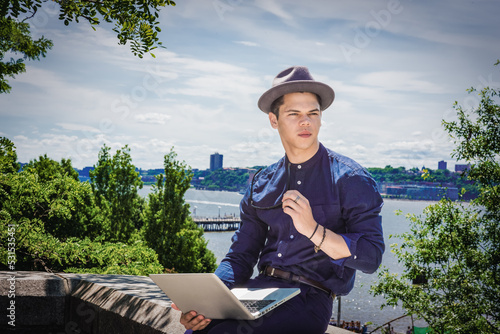 Young man, wearing blue collarless shirt, Fedora hat, hand holding sunglasses, sitting by Hudson River in New York City, reading, working on laptop computer..