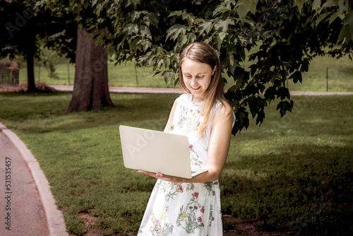 Obraz na płótnie Young college female student, wearing flower patterned white dress, standing on green lawn by trees on campus in New York City, reading, working on laptop computer. . w salonie