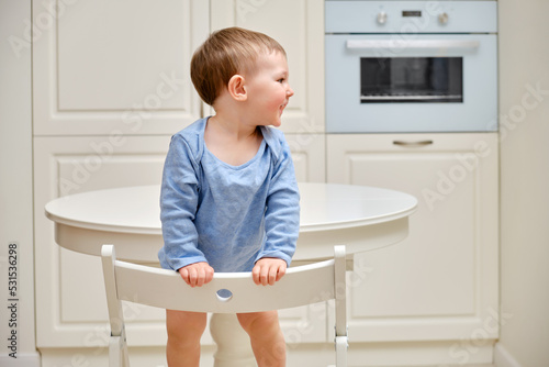 Toddler baby climbs onto a chair at the kitchen white table. A child in danger climbs up on the furniture at the risk of falling. Kid aged one year and two months