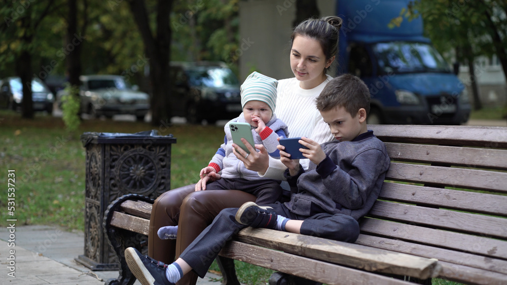 Modern family relaxing with gadgets outdoors on a bench outside. Mom and two sons in the park Modern family relaxing with gadgets outdoors on a bench outside. Mom and two sons in the park