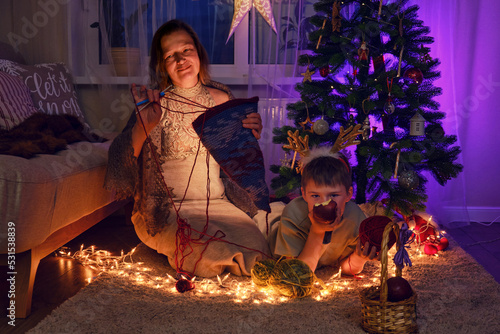 Adult woman with her son knit clothes by the Christmas tree in the evening living room. Hobbies and handicrafts with wool yarn on New Year Eve