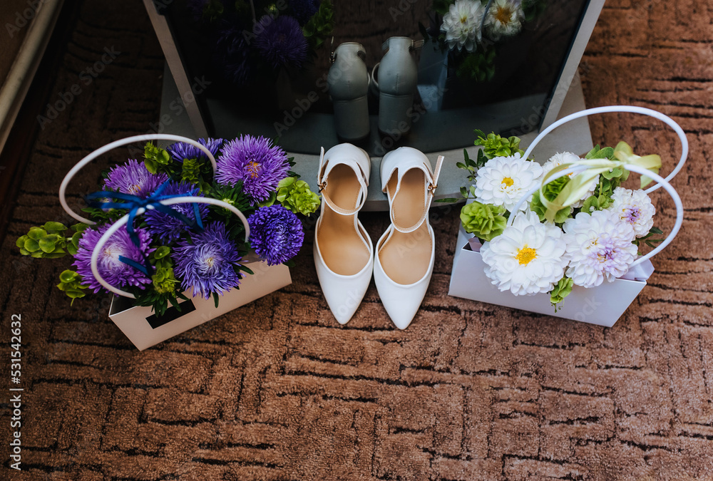 Women's white leather shoes for the bride and paper baskets with flowers stand on the floor, reflected in the mirror. Wedding photography, accessories, top view.