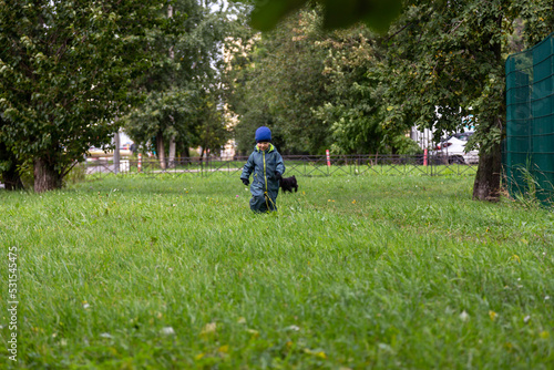 
A two-year-old boy runs through a forest park in the city on an autumn street in overalls and a hat
