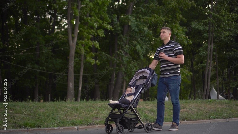 A young father carries a stroller with his son and drinks coffee in the park. A man walks with a baby in a stroller in the park.