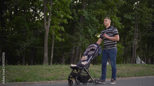 A young father carries a stroller with his son and drinks coffee in the park. A man walks with a baby in a stroller in the park.