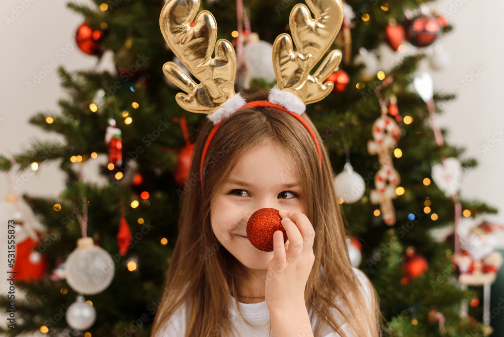 A little girl in the antlers of a Christmas deer holds a ball to her nose. Happy new year and christmas. A little girl in the antlers of a Christmas deer holds a ball to her nose. Happy new year and christmas.