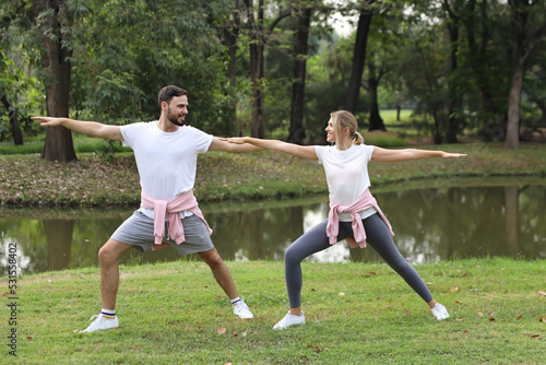 image of young caucasian couple man and woman enjoying in exercising and stretching outdoor with smiling faces and eyes contact together with green trees during summer time in the park