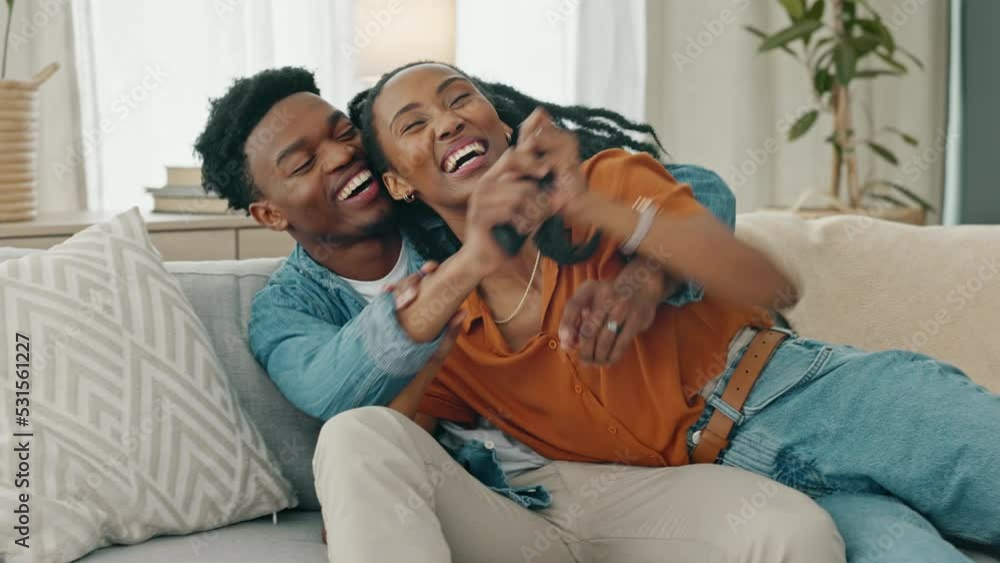Watching tv, a man and black woman with remote, playing and trying to change the channel. A happy couple cuddle on a sofa, playful and teasing while watching television in the living room at home. Watching tv, a man and black woman with remote, playing and trying to change the channel. A happy couple cuddle on a sofa, playful and teasing while watching television in the living room at home.