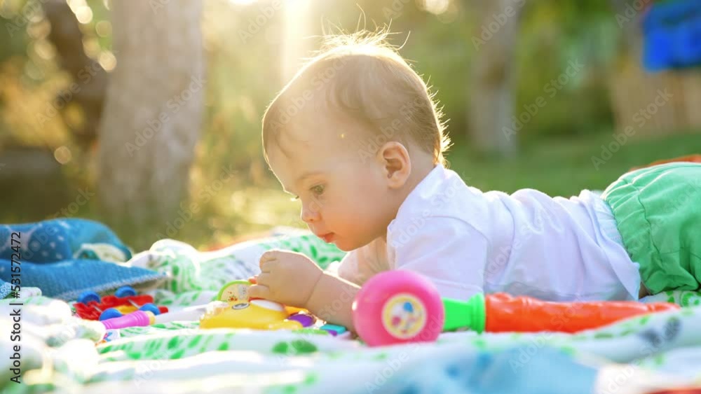 Nice little Caucasian toddler lies on his belly on the plaid in the garden. Lovely child looking at toys in the rays of bright sun. Nice little Caucasian toddler lies on his belly on the plaid in the garden. Lovely child looking at toys in the rays of bright sun.