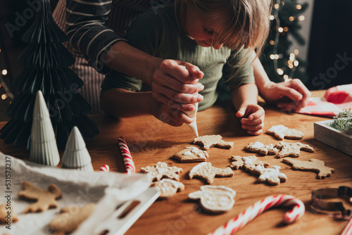 Christmas and New Year food preparation. Xmas gingerbread cooking and decorating freshly baked cookies with icing and mastic. Mom helping cute little daughter to decorate cookie on wooden messy table