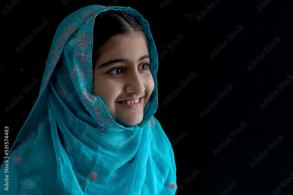 Little Muslim girl with beautiful eyes wearing blue hijab smiling with someone, black background, side view Little Muslim girl with beautiful eyes wearing blue hijab smiling with someone, black background, side view