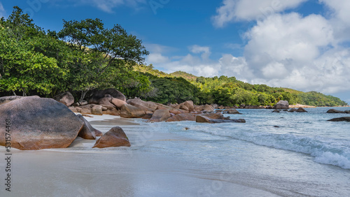 A beautiful beach on a tropical island. Picturesque granite boulders on the sand. Turquoise ocean waves are foaming. A green hill against a background of blue sky and clouds is illuminated by the sun.