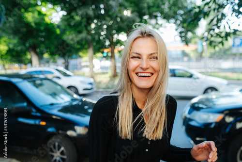 Positive young blond woman smiling in front of a car parking in background. Looking to the side. © zzzdim