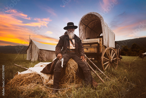 Fotografia, Obraz Oldest smart cowboy man wearing western style suite with cowboy hat holding gun on hand sit on haystack with horse carrier and tent is vintage 1800s life style concept