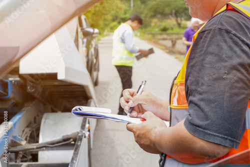 Closeup and crop insurance agent writing on clipboard while examining car after accident claim being assessed and processed on blurred damaged car truck slides with peoples and sun flare background.