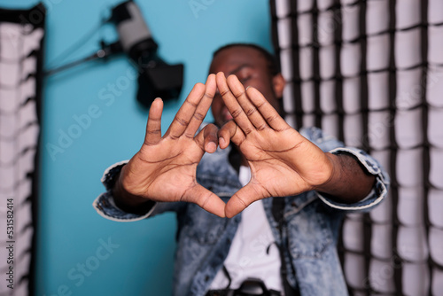 Confident producer with professional camera posing beside softboxes and spotlights. Smiling photography enthusiast making love symbol with hands while standing in production studio.