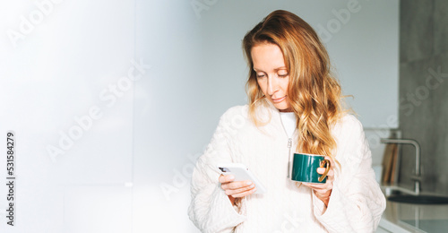 Young adult forty years beautiful blonde woman with long hair using smartphone and drinking morning coffee on kitchen at home