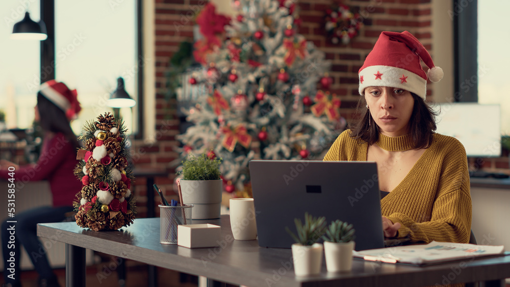 Young adult wearing santa hat and working on laptop, sending email with report in festive decorated office. Employee using computer during christmas holiday season. Tripod shot. Young adult wearing santa hat and working on laptop, sending email with report in festive decorated office. Employee using computer during christmas holiday season. Tripod shot.