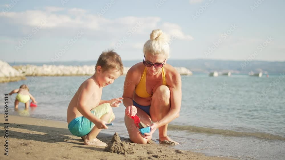 Mom plays with toys with children in the sand on the beach near the ocean. Happy family together on summer vacation spending time together in beautiful landscape. High quality 4k footage Mom plays with toys with children in the sand on the beach near the ocean. Happy family together on summer vacation spending time together in beautiful landscape. High quality 4k footage