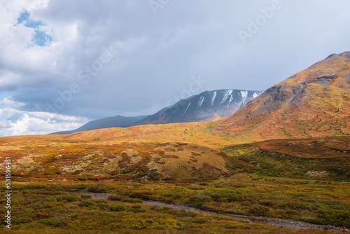 Motley autumn landscape with sunlit hills and mountain range silhouette under dramatic cloudy sky. Vivid autumn colors in mountains. Sunlight on multicolor hills and rainy clouds in changeable weather