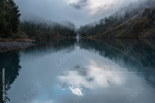 Tranquil scenery with reflex in mountain lake of snow castle in clouds. Snowy mountains in fog clearance, small river and coniferous trees reflected in alpine lake. Mountain creek and glacial lake.