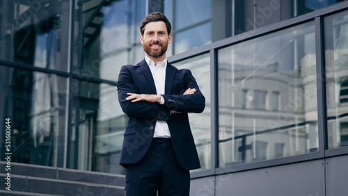 Successful confident caucasian bearded businessman in suit standing outdoors on background office building smiling proud male professional worker leader looking at camera posing with crossed arms