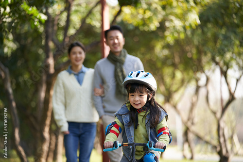 little asian girl riding bike in city park with parents in background © imtmphoto