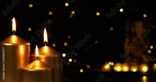 The lights of a Christmas garland glow mysteriously in the background of three burning golden candles. The flame of the candle burns steadily Concept for Christmas cards and backgrounds.