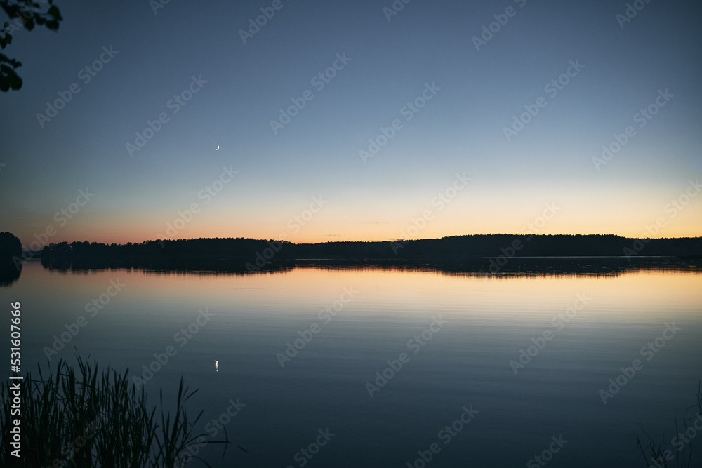 Night mystical scenery. Crescent moon over the misty lake with forest on the other coast and its reflection in the still water.