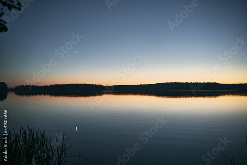 Night mystical scenery. Crescent moon over the misty lake with forest on the other coast and its reflection in the still water.