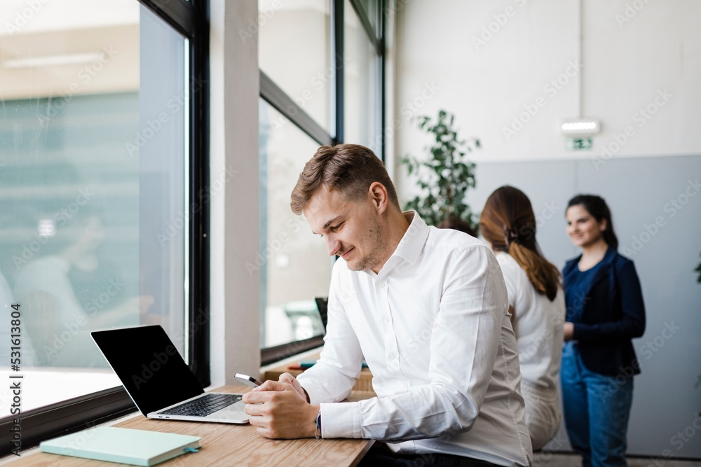 Smiling businessman using mobile phone sitting by laptop in office Smiling businessman using mobile phone sitting by laptop in office