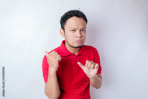 Upset Asian man wearing a red t-shirt pointing at the copy space beside him, isolated by a white background