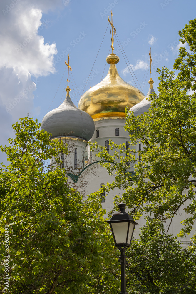 Golden and gray domes of the Orthodox Church with holy crosses, greenery and a lantern. Novodevichy Convent in Moscow.  Cloudy sky on a summer day.