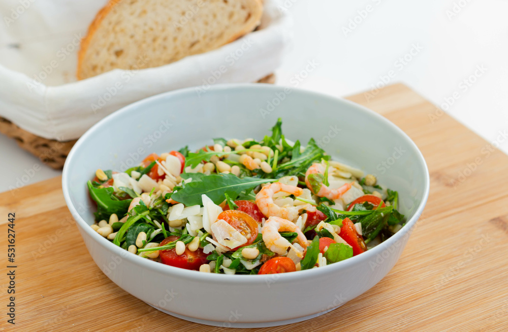 Salad with shrimps, arugula, cherry tomatoes, pine nuts, cheese in a white bowl on a wooden board.