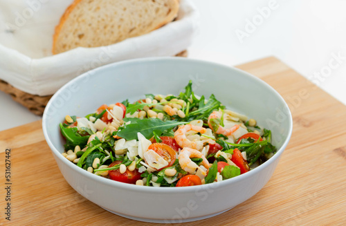 Salad with shrimps, arugula, cherry tomatoes, pine nuts, cheese in a white bowl on a wooden board.