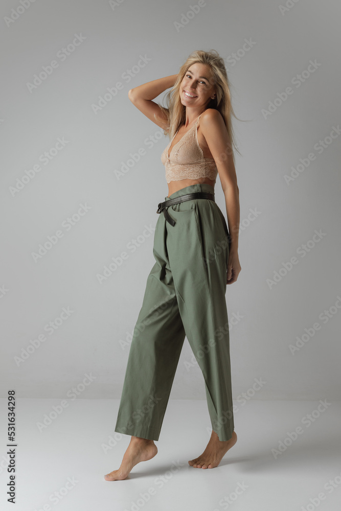 Elegance. Young woman with long straight blonde hair and natural makeup wearing crop top and casual trousers posing over grey background.