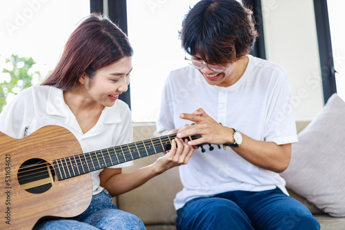Happy couple sitting on couch and young man teaching girlfriend playing guitar in living room