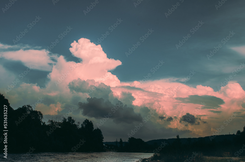Obraz premium Beautiful cloudy sky at sunset over the river. The high bank of the river is overgrown with grass.