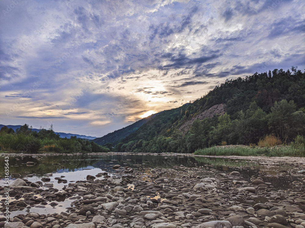 Fototapeta premium Beautiful cloudy sky at sunset over the river. The high bank of the river is overgrown with grass.
