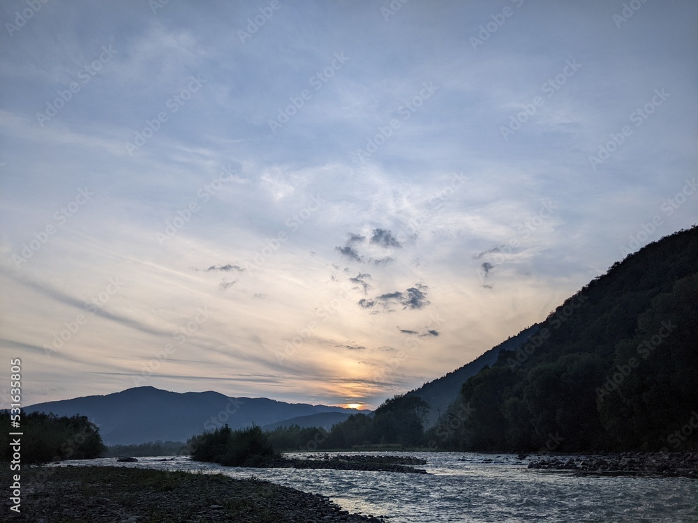 Beautiful cloudy sky at sunset over the river. The high bank of the river is overgrown with grass.