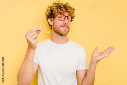 Young caucasian man wearing hearing aid isolated on yellow background showing a copy space on a palm and holding another hand on waist.