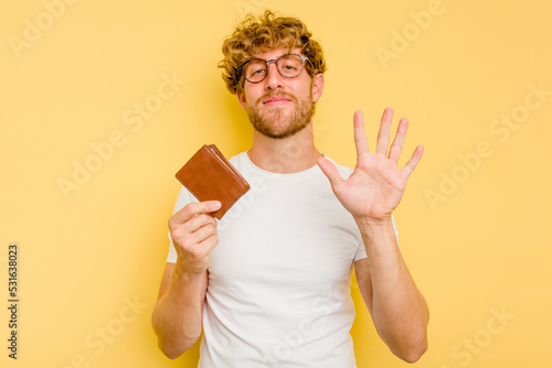 Young caucasian man holding a wallet isolated on yellow background smiling cheerful showing number five with fingers.