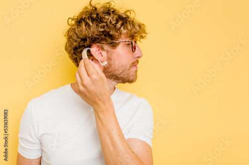 Young caucasian man wearing hearing aid isolated on yellow background