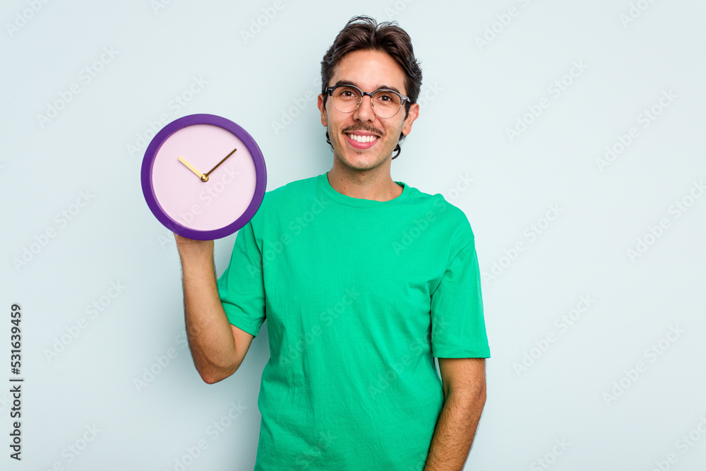 Young hispanic man holding a clock isolated on white background happy, smiling and cheerful. Young hispanic man holding a clock isolated on white background happy, smiling and cheerful.