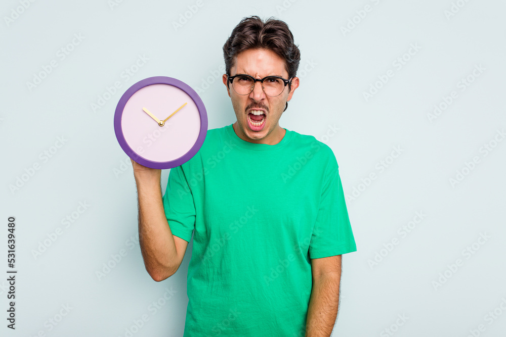 Young hispanic man holding a clock isolated on white background screaming very angry and aggressive.