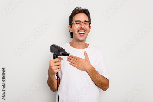 Young hispanic man holding a hairdryer isolated on white background laughs out loudly keeping hand on chest.